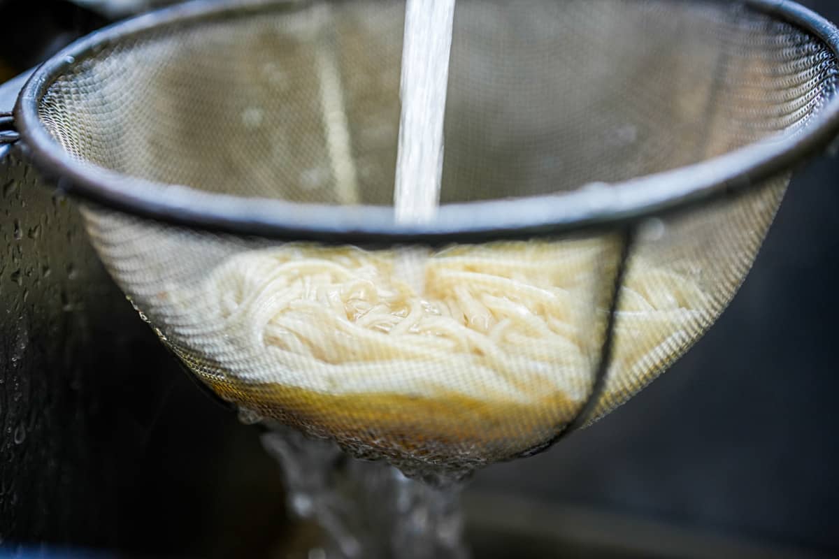 Noodles being rinsed in a mesh strainer under running water.