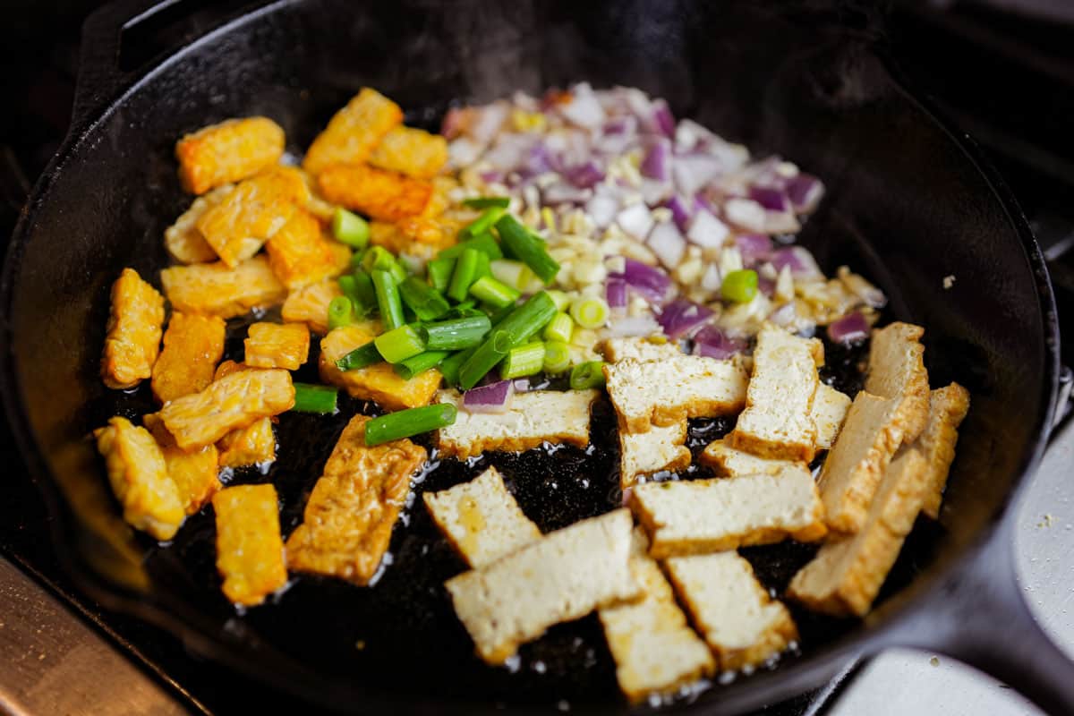 tempeh, tofu, red onions, garlic and scallions being stir fried on the stove top.