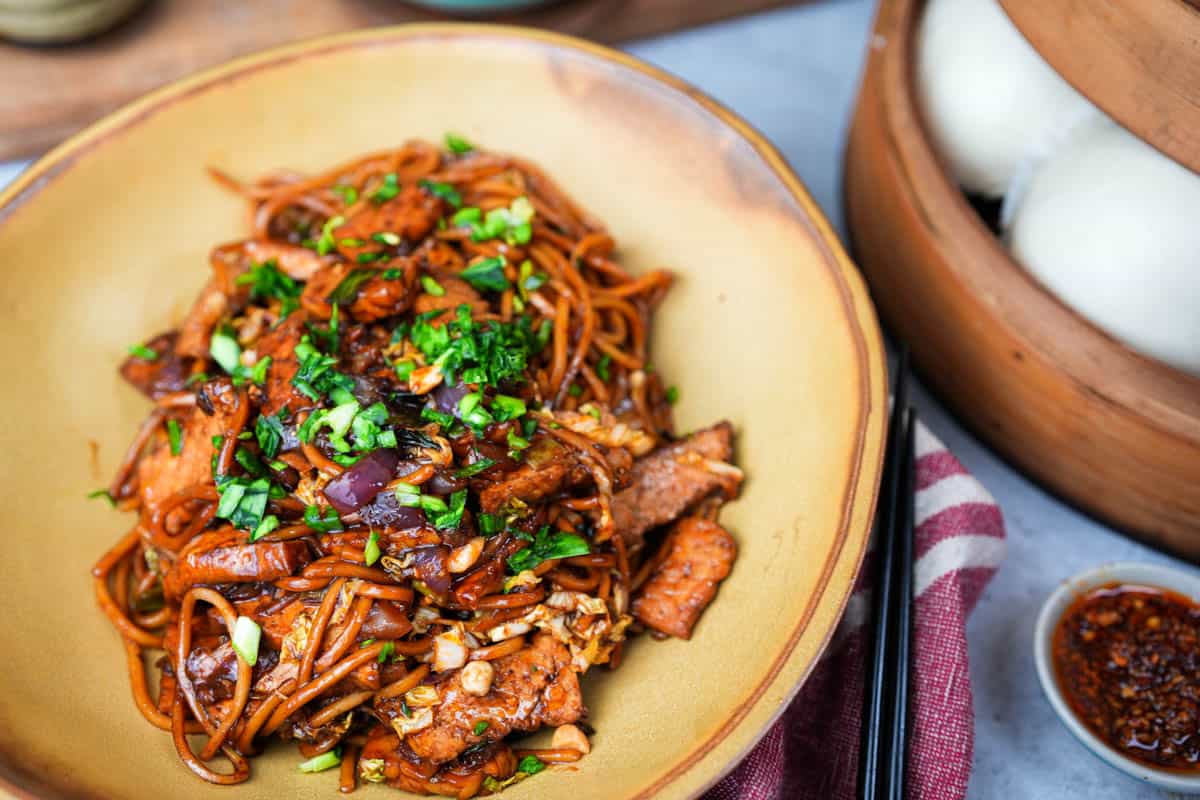 Mie goreng in a brown bowl with chopsticks, chili crisp, and a tray of steamed buns on the side.