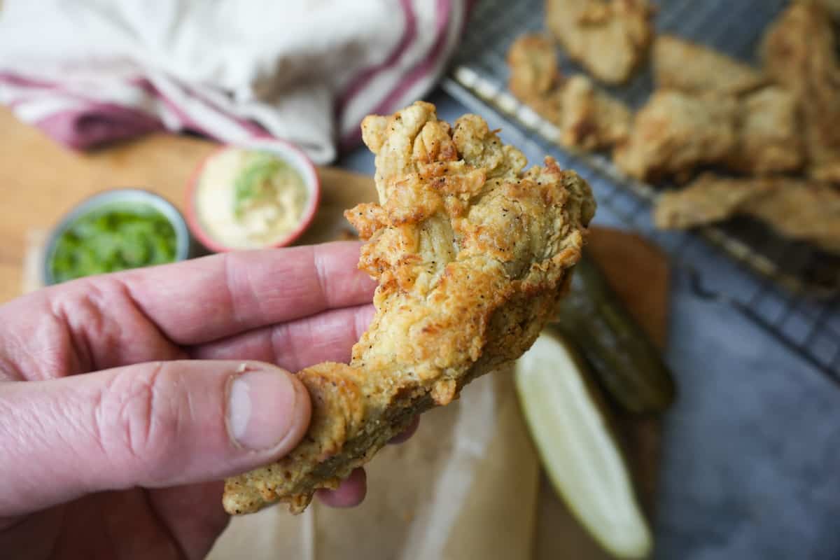 A single handheld piece of fried seitan. An out of focus tray of vegan fried chicken and dips in the background.