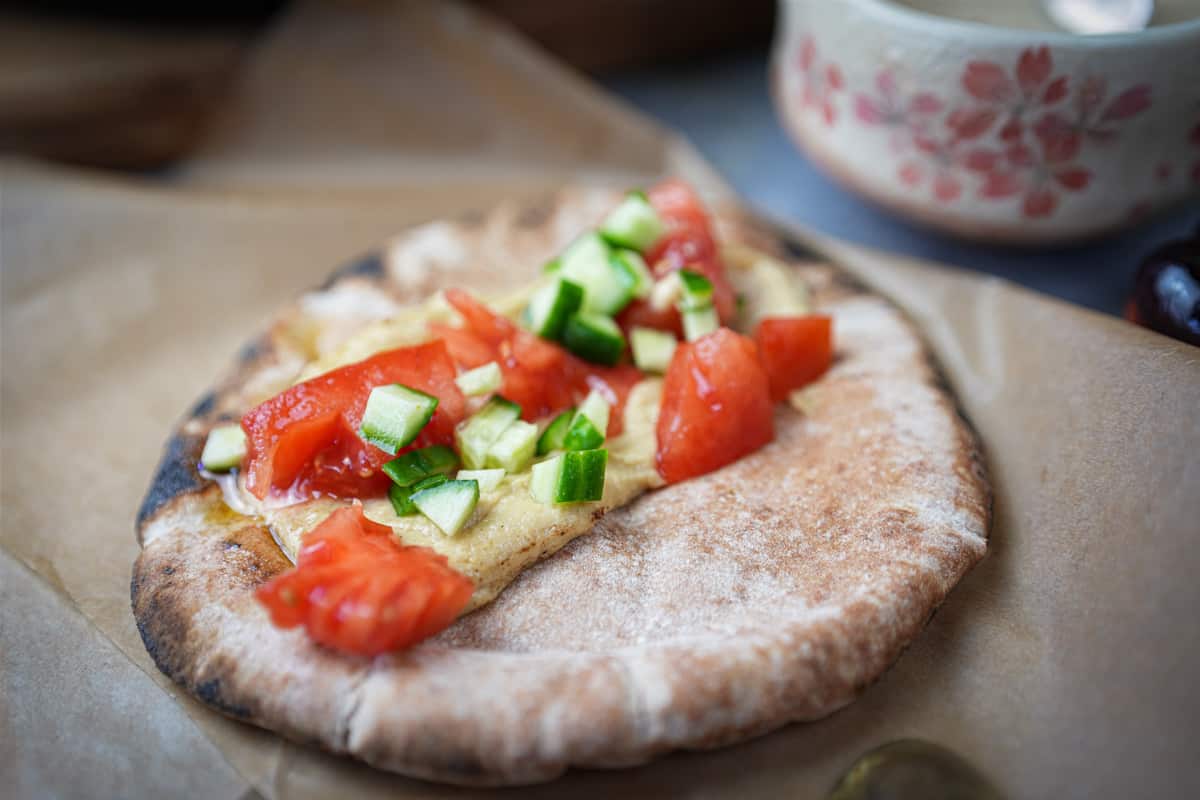 tomatoes and cucumbers being put onto a grilled pita with hummus.