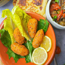 Kofte in an orange bowl with lettuce leave and lemon slices. Servings of labneh, msemen, irmik helvasi, loubia, and turkish Shakshuka are on the side.