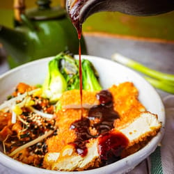A bowl of tofu katsu with sauce being poured over it.