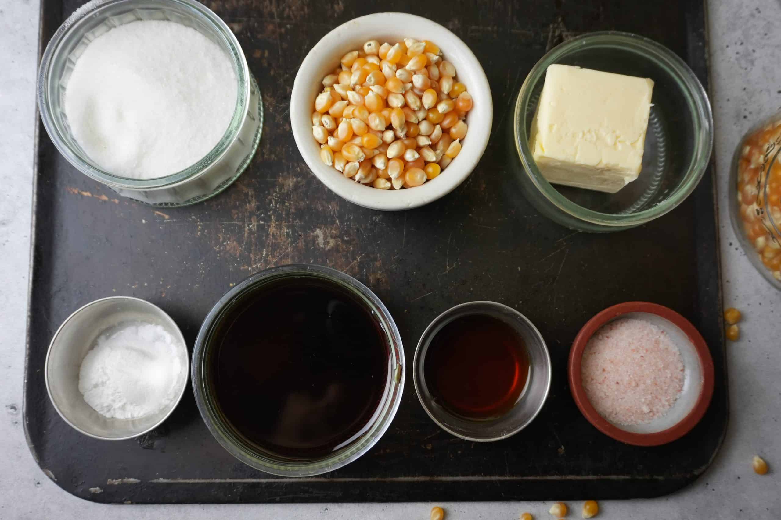 The ingredients for vegan caramel popcorn in small cups on a black metal tray- sugar, popcorn, salt, vegan butter, vanilla extract, baking soda, and maple syrup