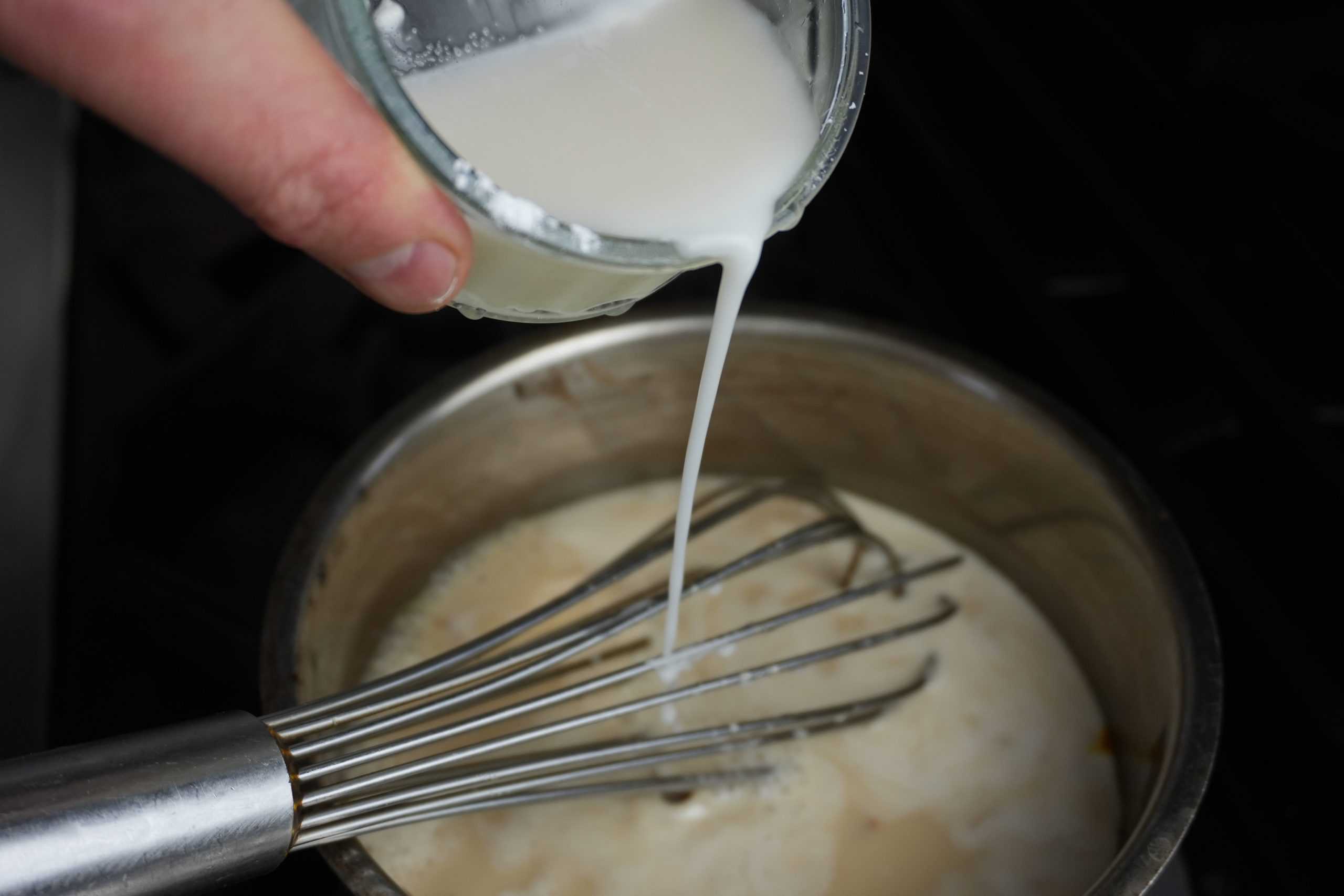 horozontal shot showing slurring being poured into a pot of custard to thicken it. Pot has a whisk in it.