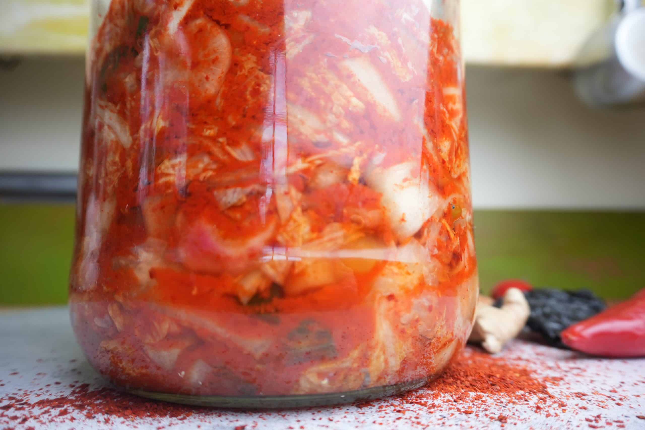 fermented kimchi in a glass jar with kimchi ingredients on the counter around it.