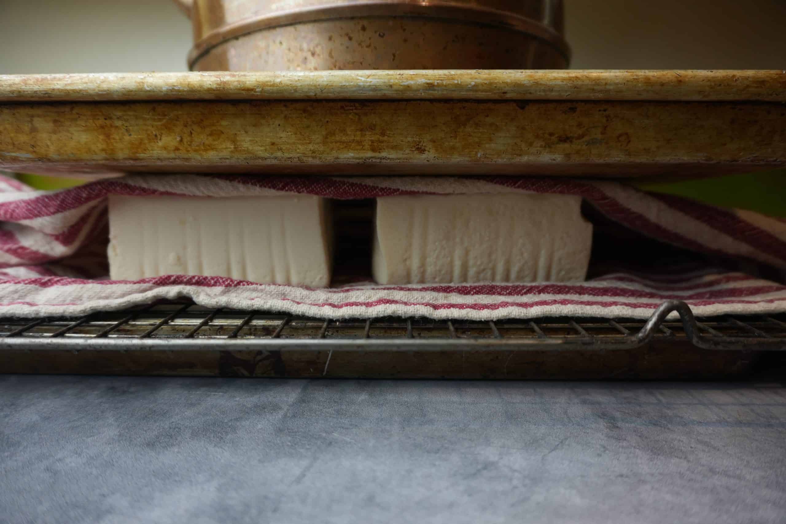 Soft tofu being pressed. it is wrapped in a towel on a cooling rack with a heavy pan on top of it.