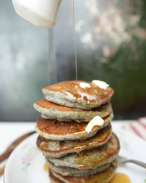 blue corn pancakes with pine nut butter in a dish. Maple syrup being poured on them from above.