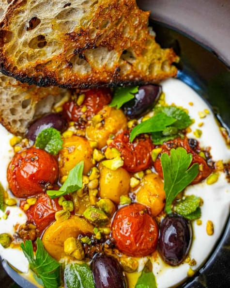 Labneh in a black bowl topped with roasted tomatoes, herbs and pistachios. Grilled bread on the side.