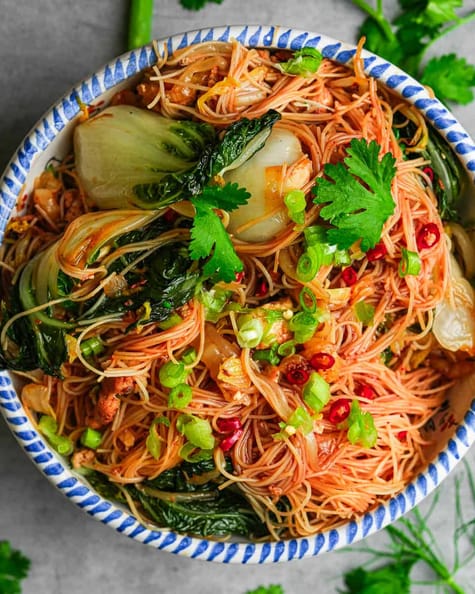 A ceramic bowl of noodles with vegetables and herbs.