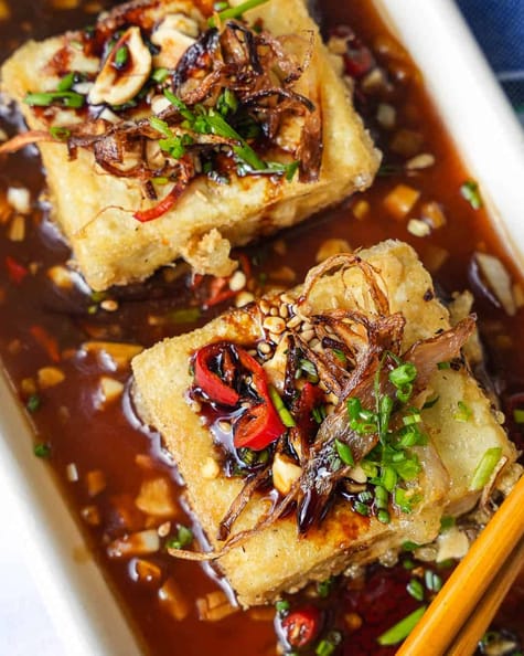 Two squares of tofu in sauce with chopsticks.