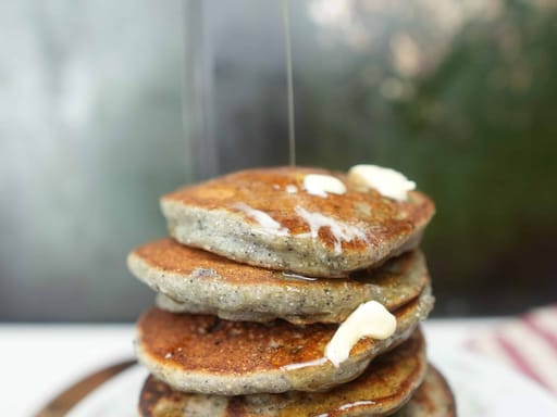 blue corn pancakes with pine nut butter in a dish. Maple syrup being poured on them from above.