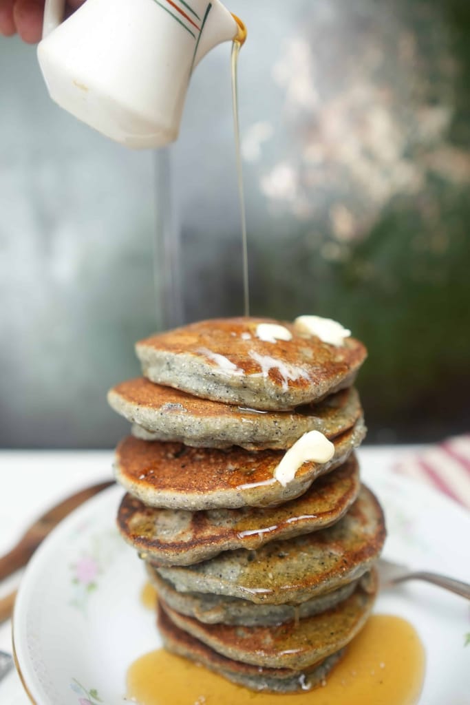 Blue corn pancakes with pine nut butter in a dish getting maple syrup poured on them from a tiny cute pitcher 