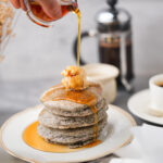 A hand pouring syrup onto a stack of blue corn pancakes topped with butter, with a French press, coffee cup, and other breakfast items in the background.