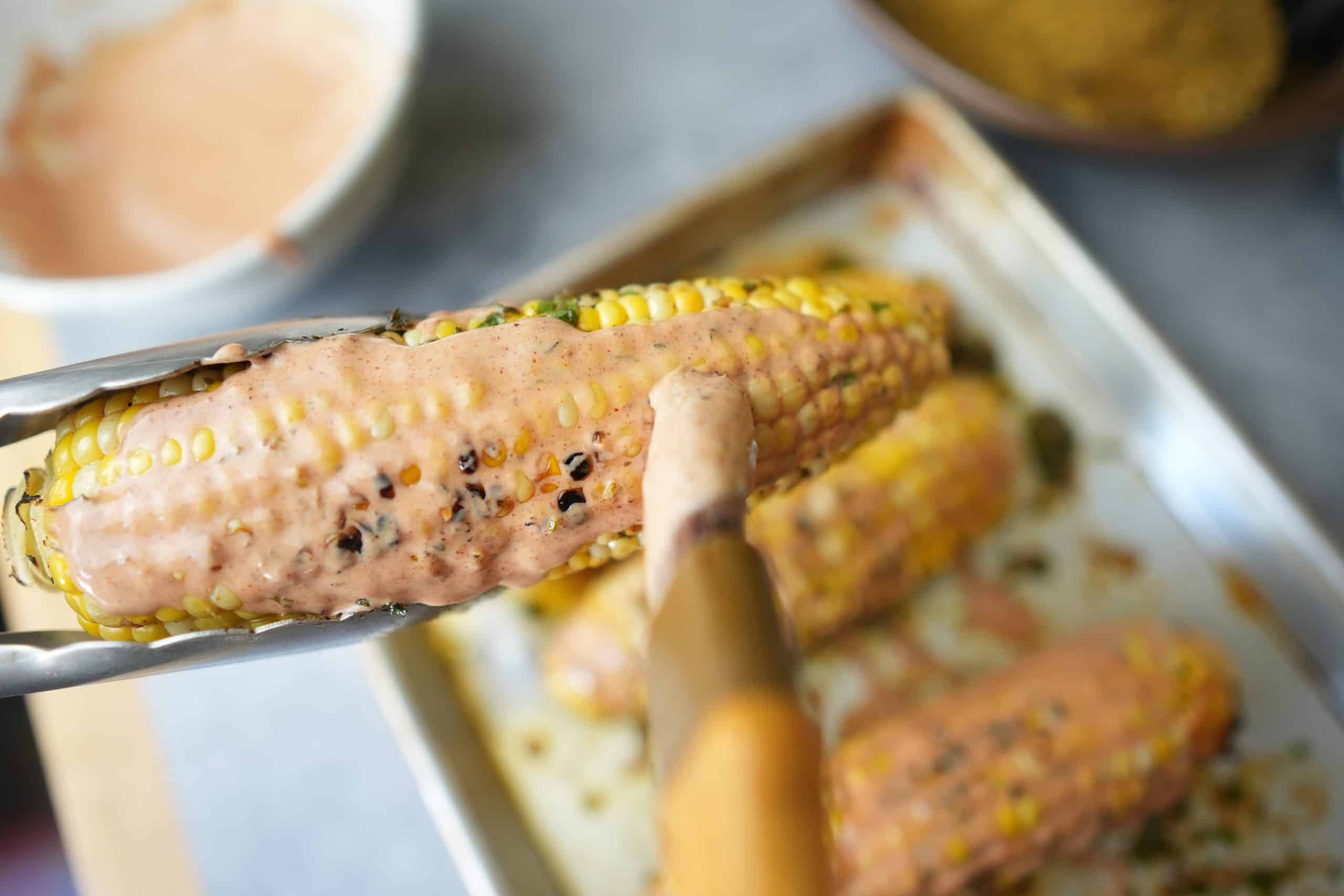 ancho chili mayo being put onto the grilled corn with a pastry brush.