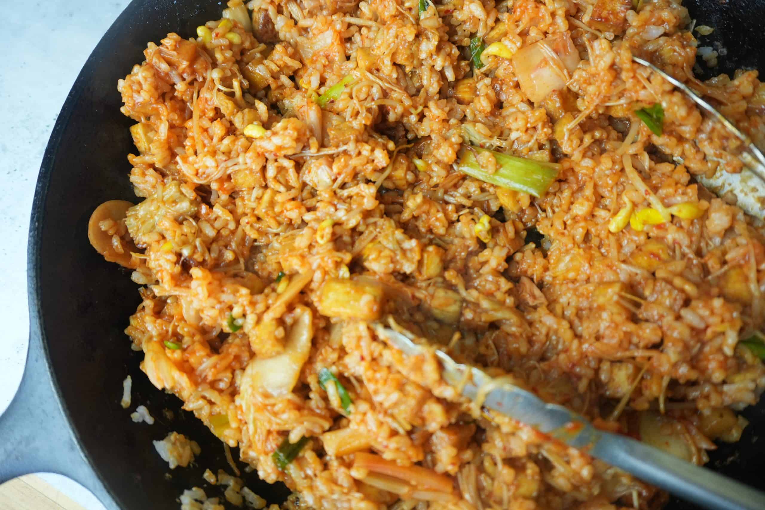 All of the ingredients for vegan kimchi fried rice stir frying together in a large cast iron skillet. Tongs in the pan