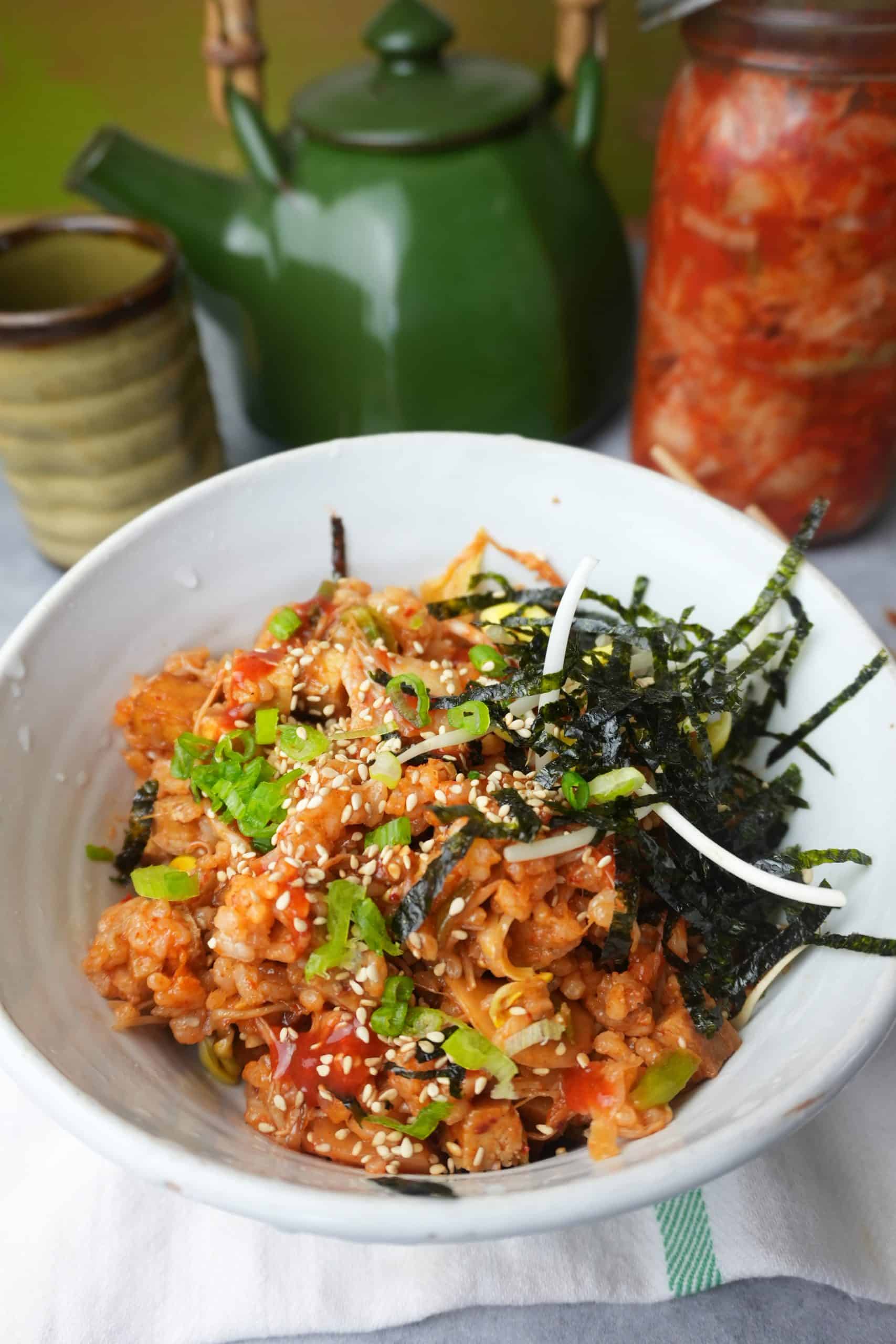 vegan kimchi fried rice in a white bowl. Napkin underneath the bowl. Teacup and pitcher and a jar of kimchi in the background