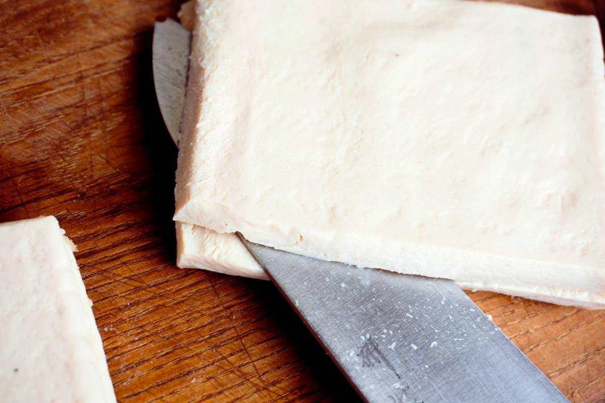 a serving of pressed tofu being cut in half on a wooden surface with a Japanese knife