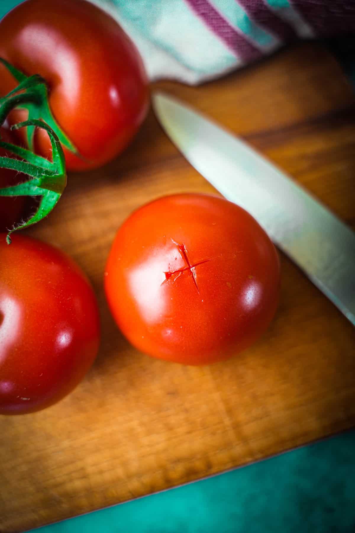 An X is cut into the bottom of a tomato before blanching. A knife and more tomatoes are in the background.