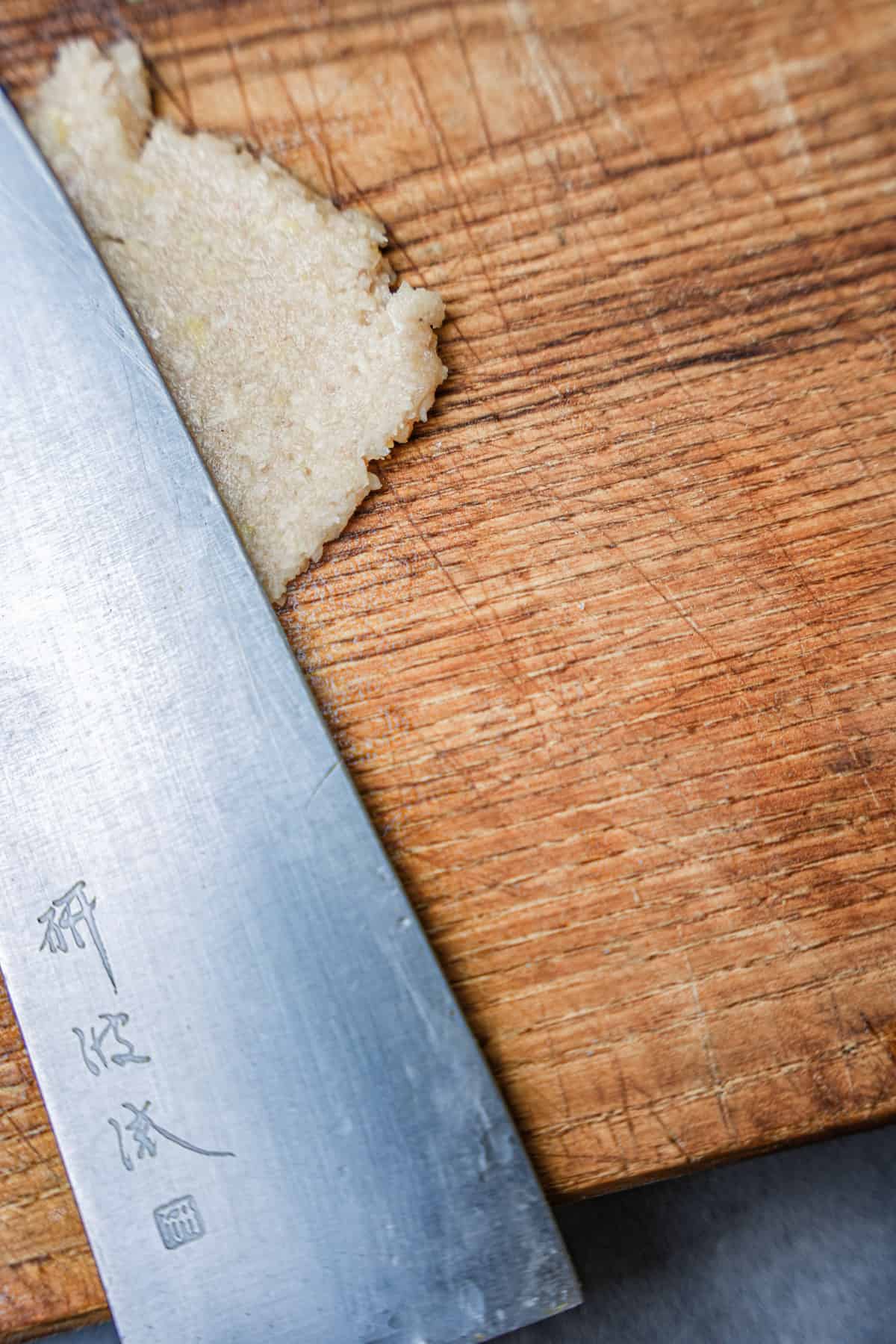 Garlic paste is pressed by the side of a knife on a wooden cutting board.