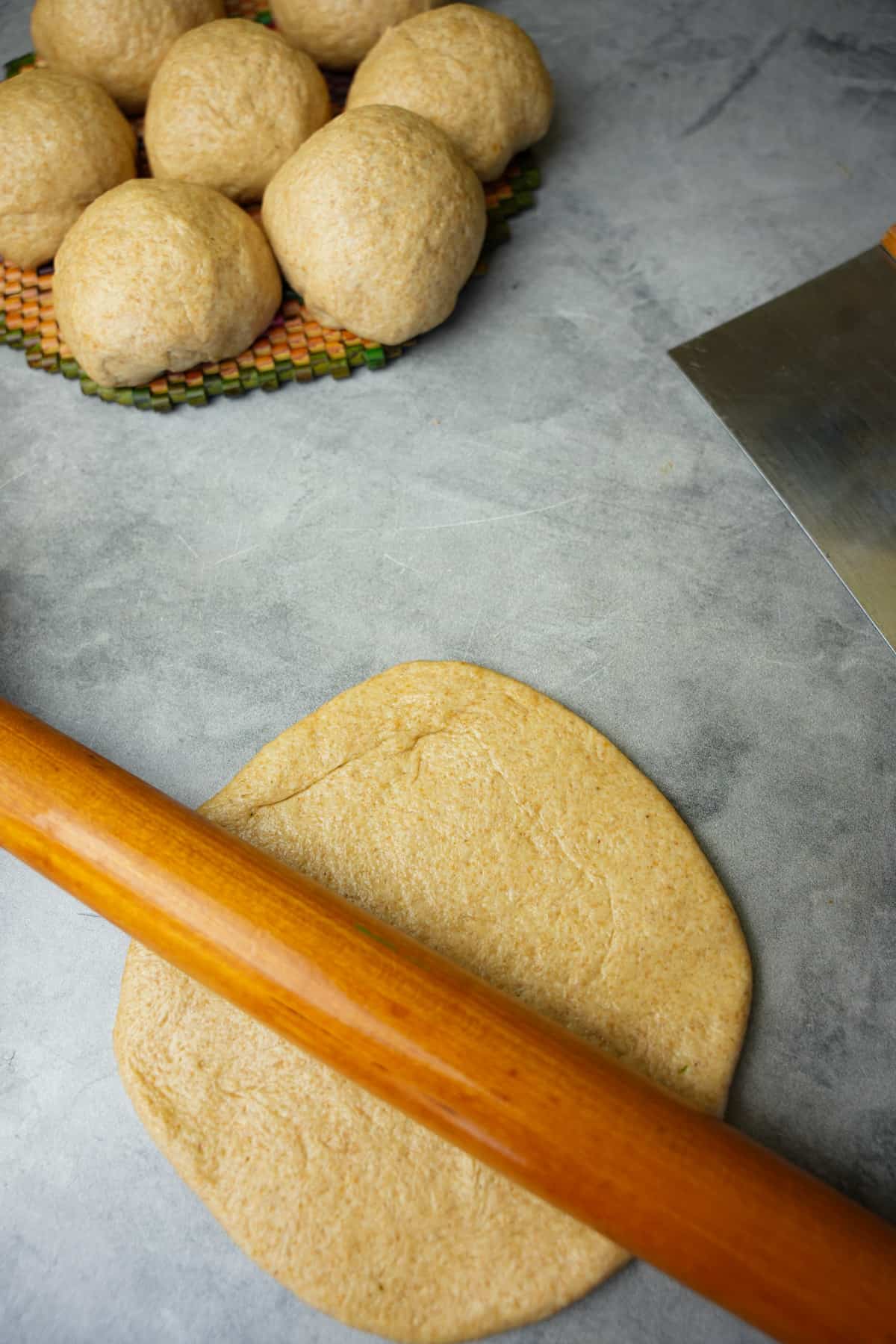 Rolling out a single portion of dough with a wooden rolling pin. Additional rounded dough can be seen in the background on a mat.