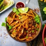 A handpainted small bowl filled with bami goreng garnished with scallions, cilantro, and a lime wedge. Chili sauce and steamed bok choy on the side.