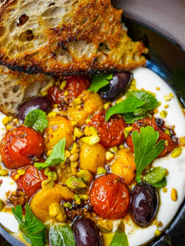 Labneh in a black bowl topped with roasted tomatoes, herbs and pistachios. Grilled bread on the side.