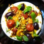 Labneh in a black bowl topped with roasted tomatoes, herbs and pistachios.