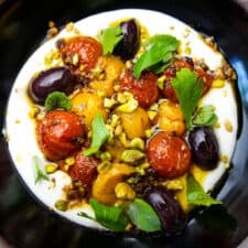 Labneh in a black bowl topped with roasted tomatoes, herbs and pistachios.