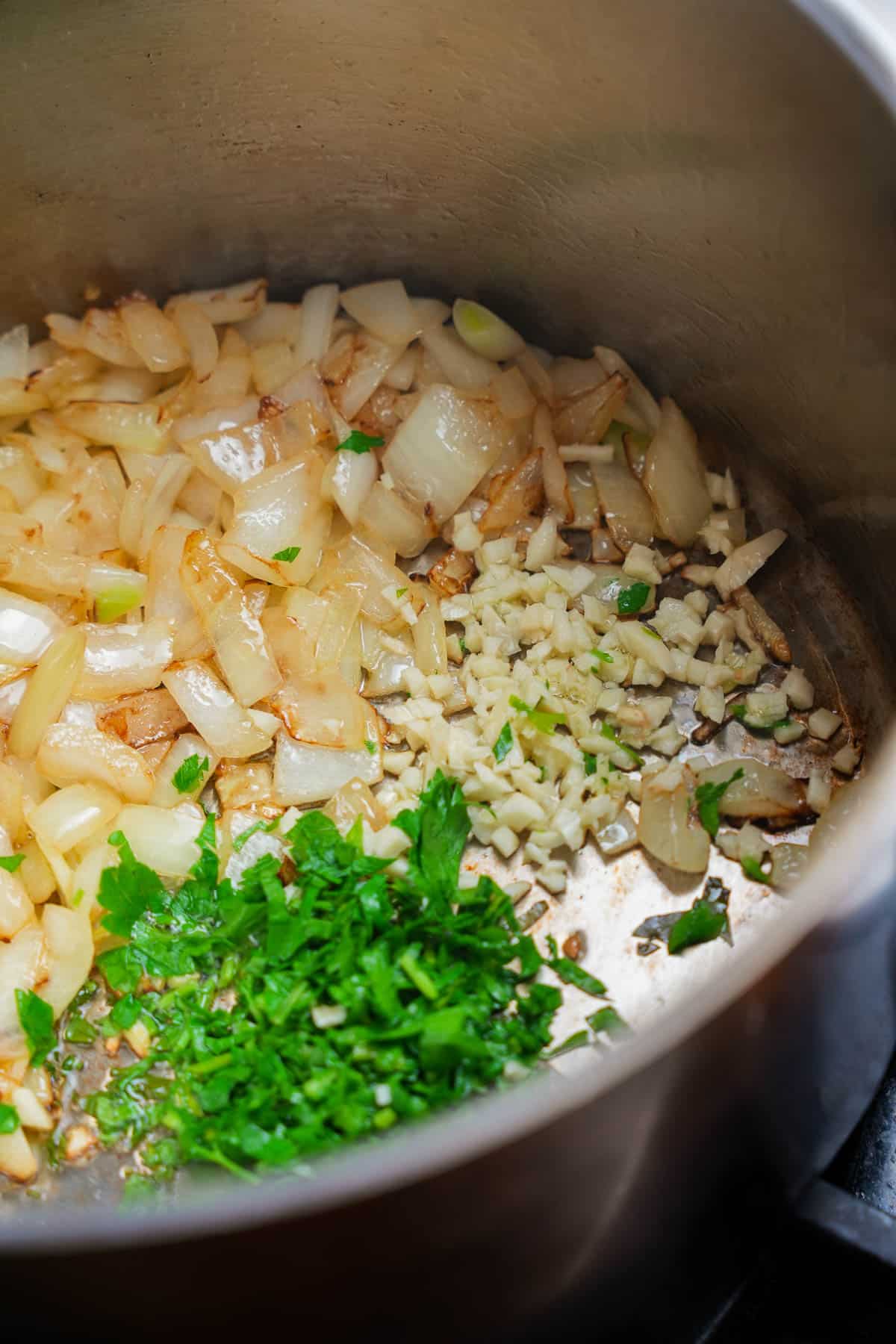 Garlic and parsley are added to the cooking onions in a stainless steel pot.