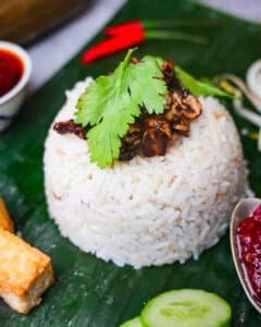 Nasi Uduk betawi topped with seasoned roasted peanuts and cilantro on a banana leaf with tofu, cucumbers, sambal and other sides.