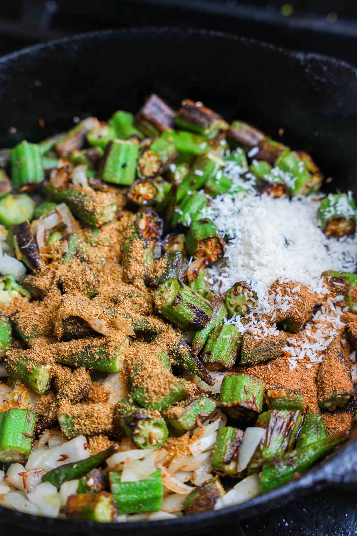 Spices and coconut are added to the pan frying okra in a cast iron skillet.
