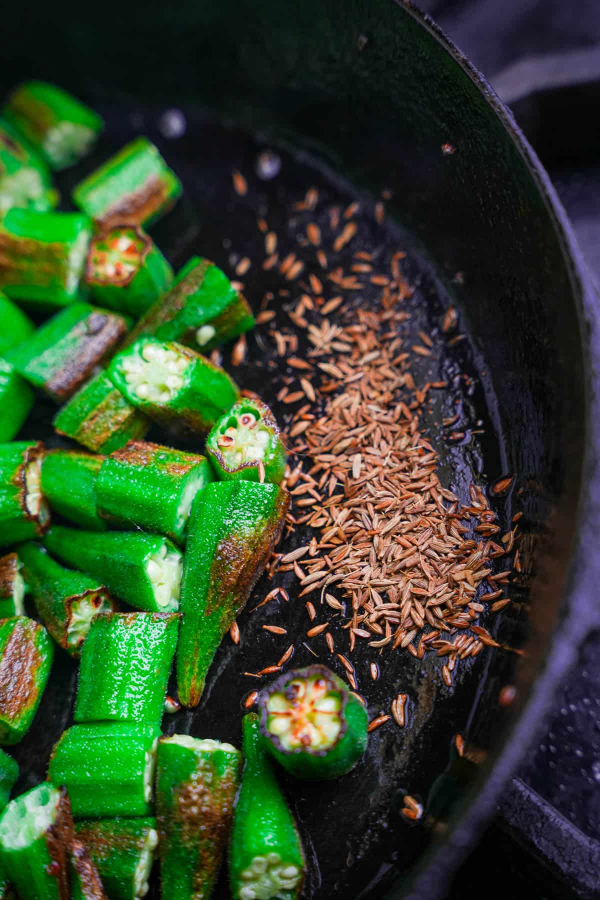 Cumin seeds are added directly to the bare metal surface of a cast iron pan next to the cooking okra.