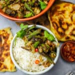 Punjabi dry bhindi over steamed basmati rice in a white bowl garnished with sliced red chilies and cilantro. A fork, mango pickle, and freshly grilled Indian bread are on the side.