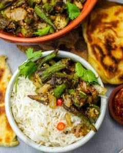 Punjabi dry bhindi over steamed basmati rice in a white bowl garnished with sliced red chilies and cilantro. A fork, mango pickle, and freshly grilled Indian bread are on the side.