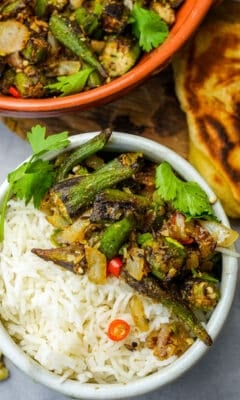 Punjabi dry bhindi over steamed basmati rice in a white bowl garnished with sliced red chilies and cilantro. A fork, mango pickle, and freshly grilled Indian bread are on the side.