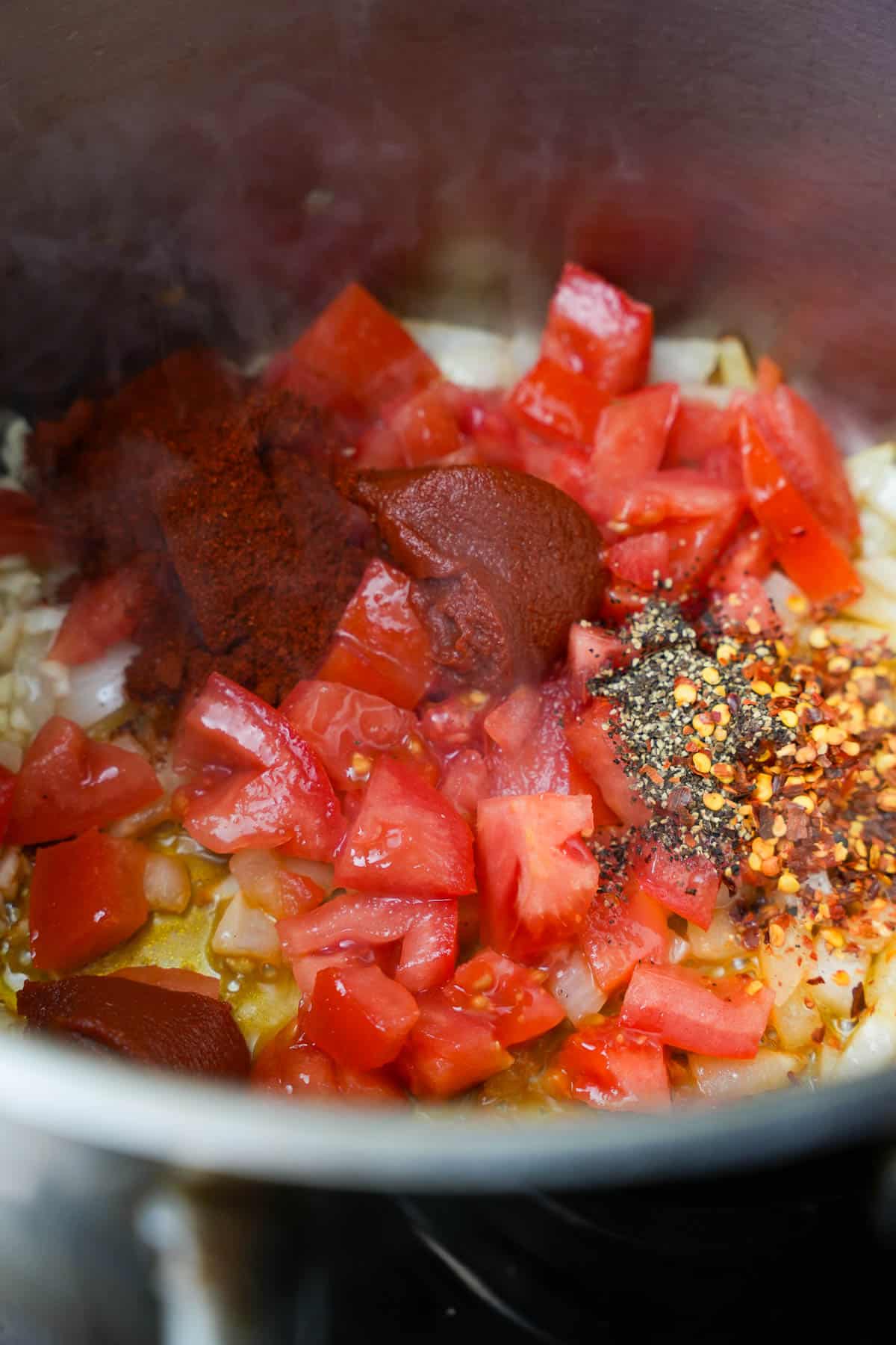 tomatoes and seasonings are added to the cooking onions in a stainless steel pot.