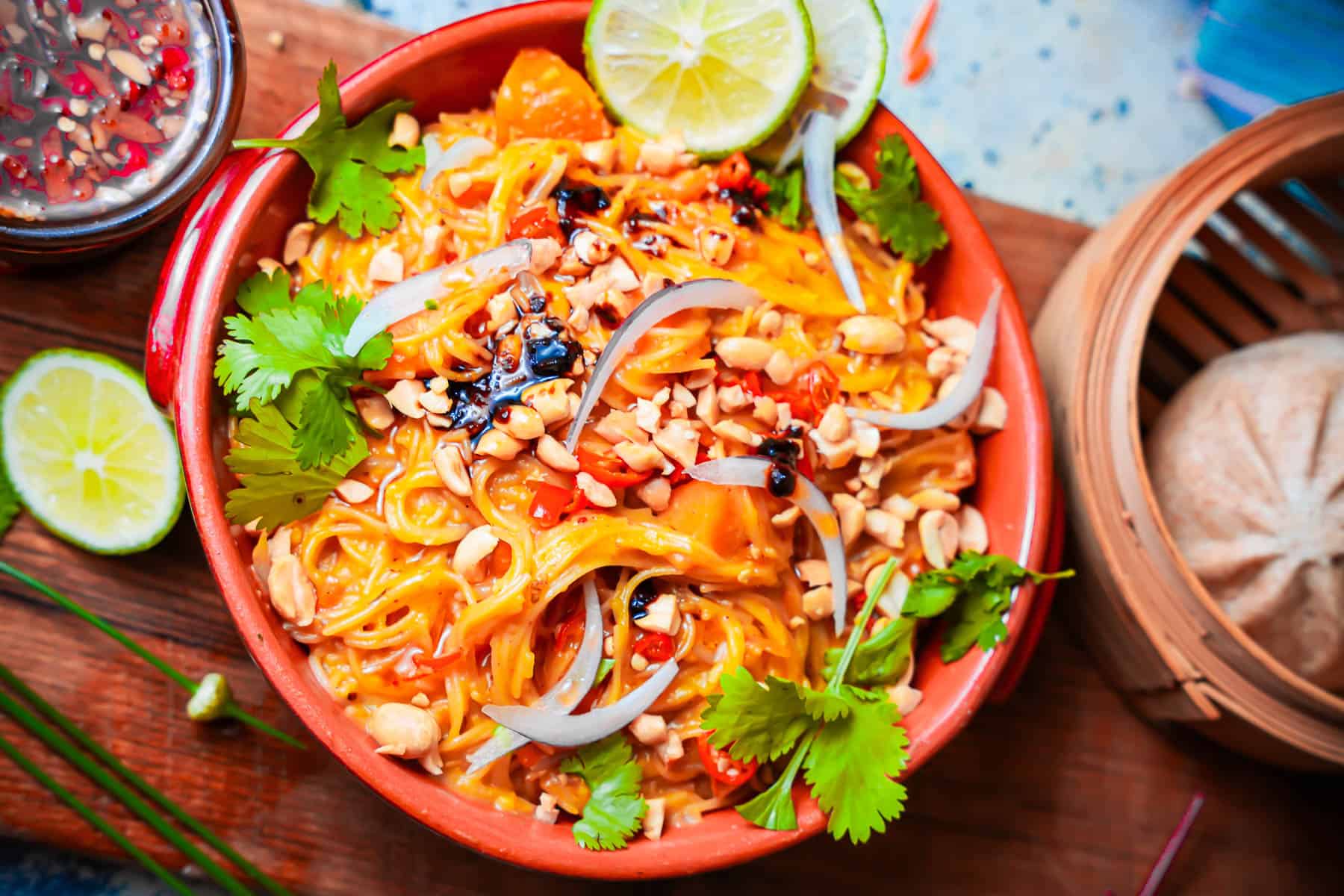 Khao suey in an orange bowl garnished with thinly sliced red onions, chili oil, peanuts, cilantro and lime leaves.