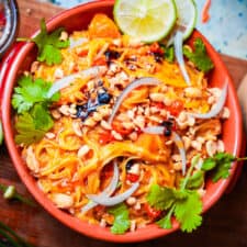 Khao suey in an orange bowl garnished with thinly sliced red onions, chili oil, peanuts, cilantro and lime leaves.