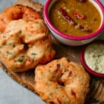 Medhu vadai on a wooden board with sambar, coconut chutney and a chili pepper.