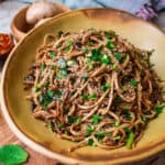 A brown handmade wide bowl with Mi Xao in it, garnished with sesame seeds and fresh chives.