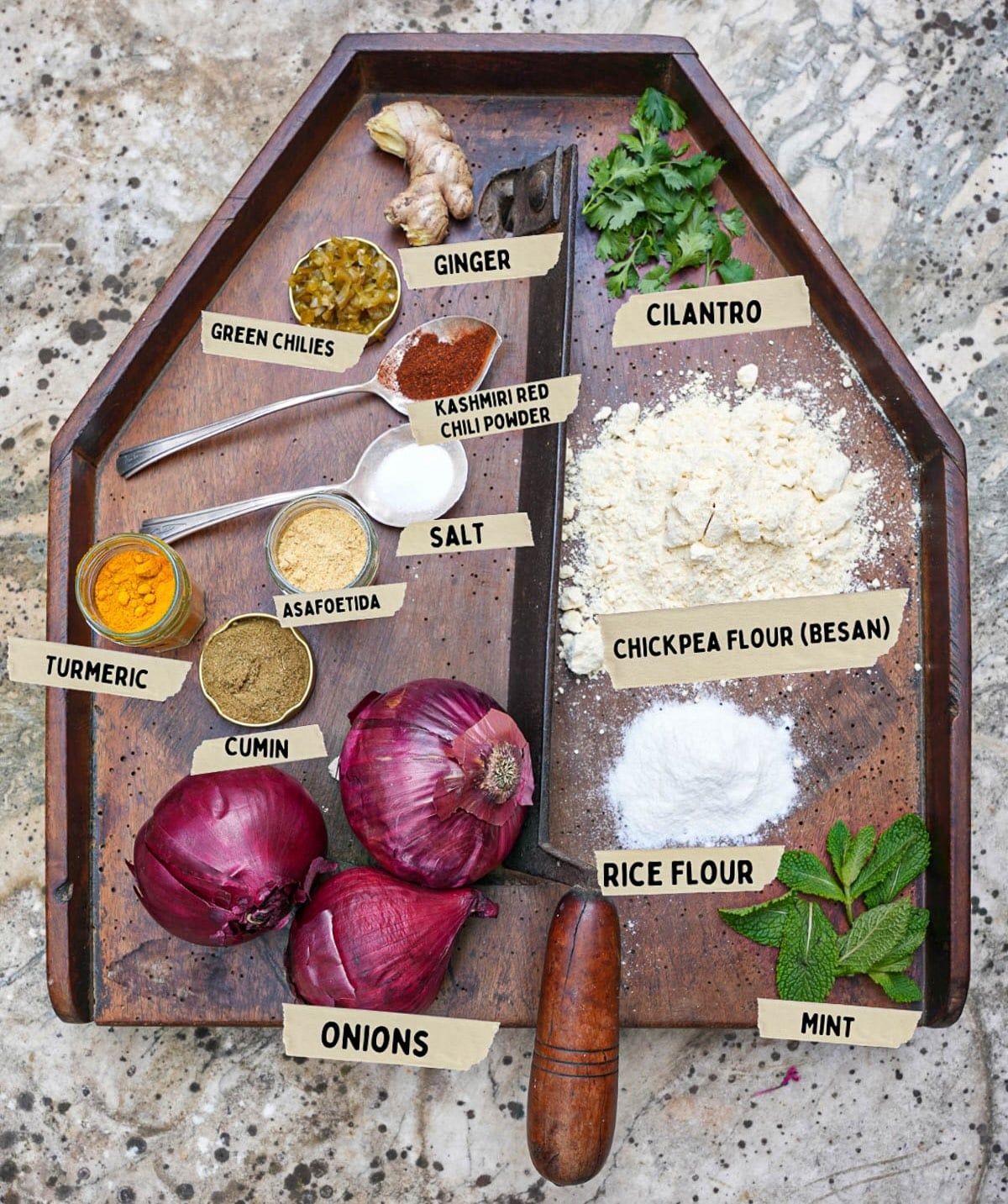 A wooden tray with labeled spices and ingredients: ginger, cilantro, green chiles, red chili powder, salt, chickpea flour for bhaji, rice flour, turmeric, asafoetida, cumin, onions,and mint.