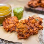 Three nion bhaji on a piece of craft paper with a wooden board in the background with more bhaji. Side cups next to the bhaji contain mint yogurt sauce and mango chutney.