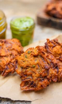 Three nion bhaji on a piece of craft paper with a wooden board in the background with more bhaji. Side cups next to the bhaji contain mint yogurt sauce and mango chutney.