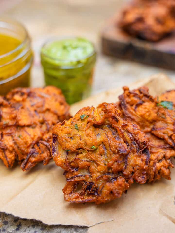 Three nion bhaji on a piece of craft paper with a wooden board in the background with more bhaji. Side cups next to the bhaji contain mint yogurt sauce and mango chutney.