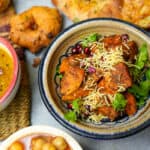 Shakarkandi chaat in a colorful ceramic bowl, garnished with pomegranate seeds, sev and cilantro. Channa, vadai, and kulcha in the background.