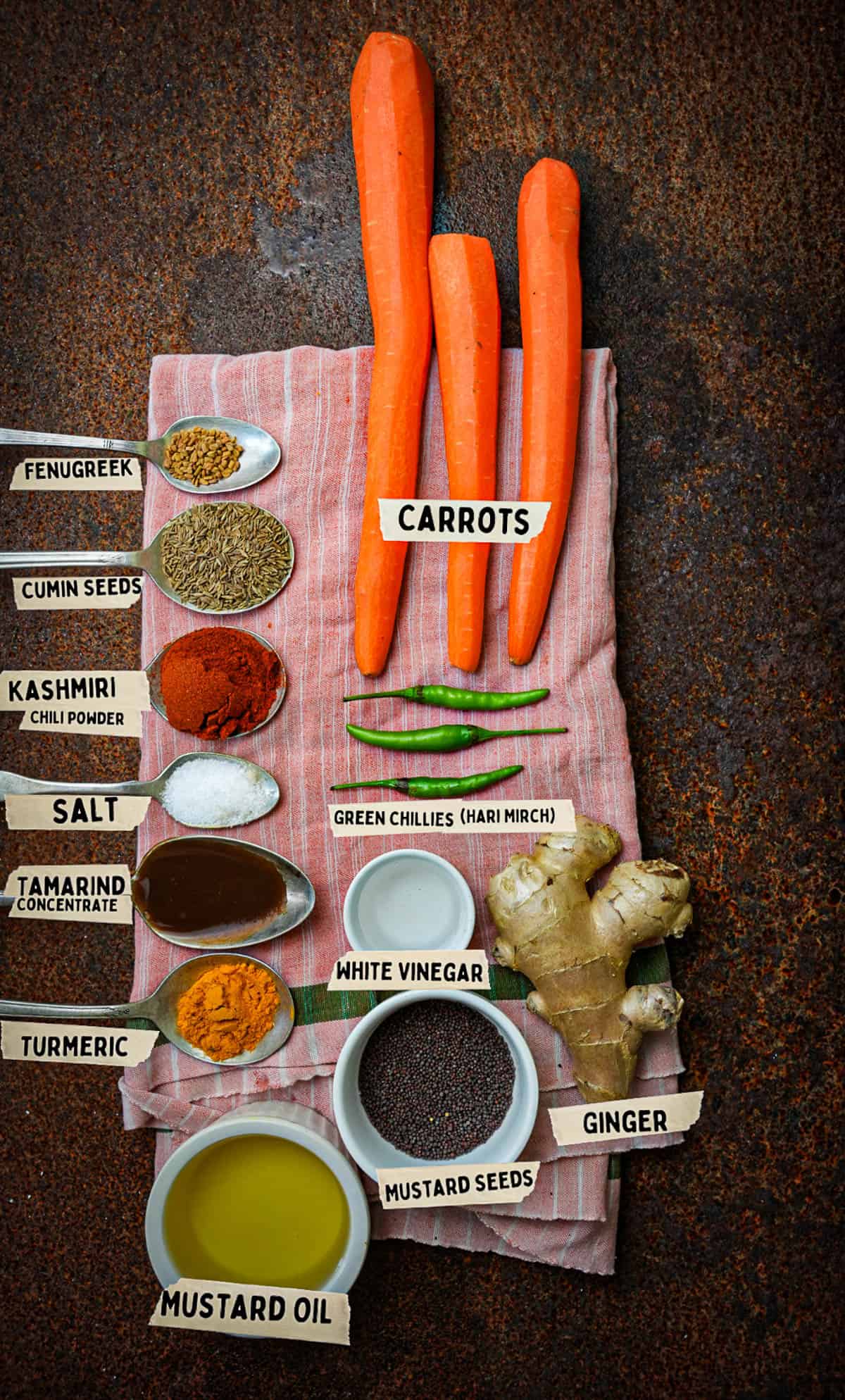 carrot pickle ingredients measured and labeled on a pink cloth on top of a rusted metal surface.