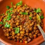 chickpeas garnished with cilantro and chilies in an orange ceramic bowl with a spoon.