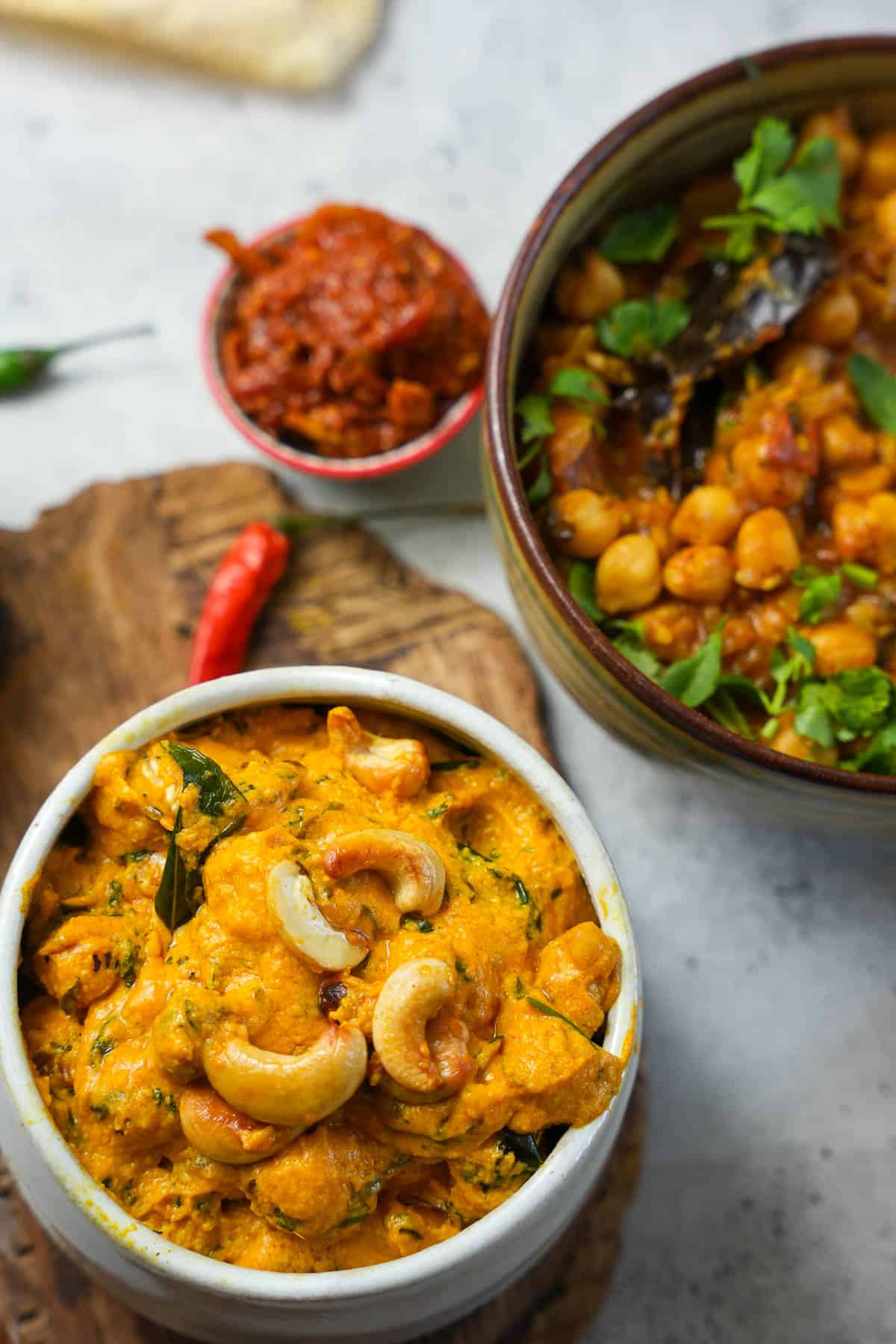 A white ceramic bowl of phool makhana curry with cashews. A bowl of chana masala and mango pickle in the background.