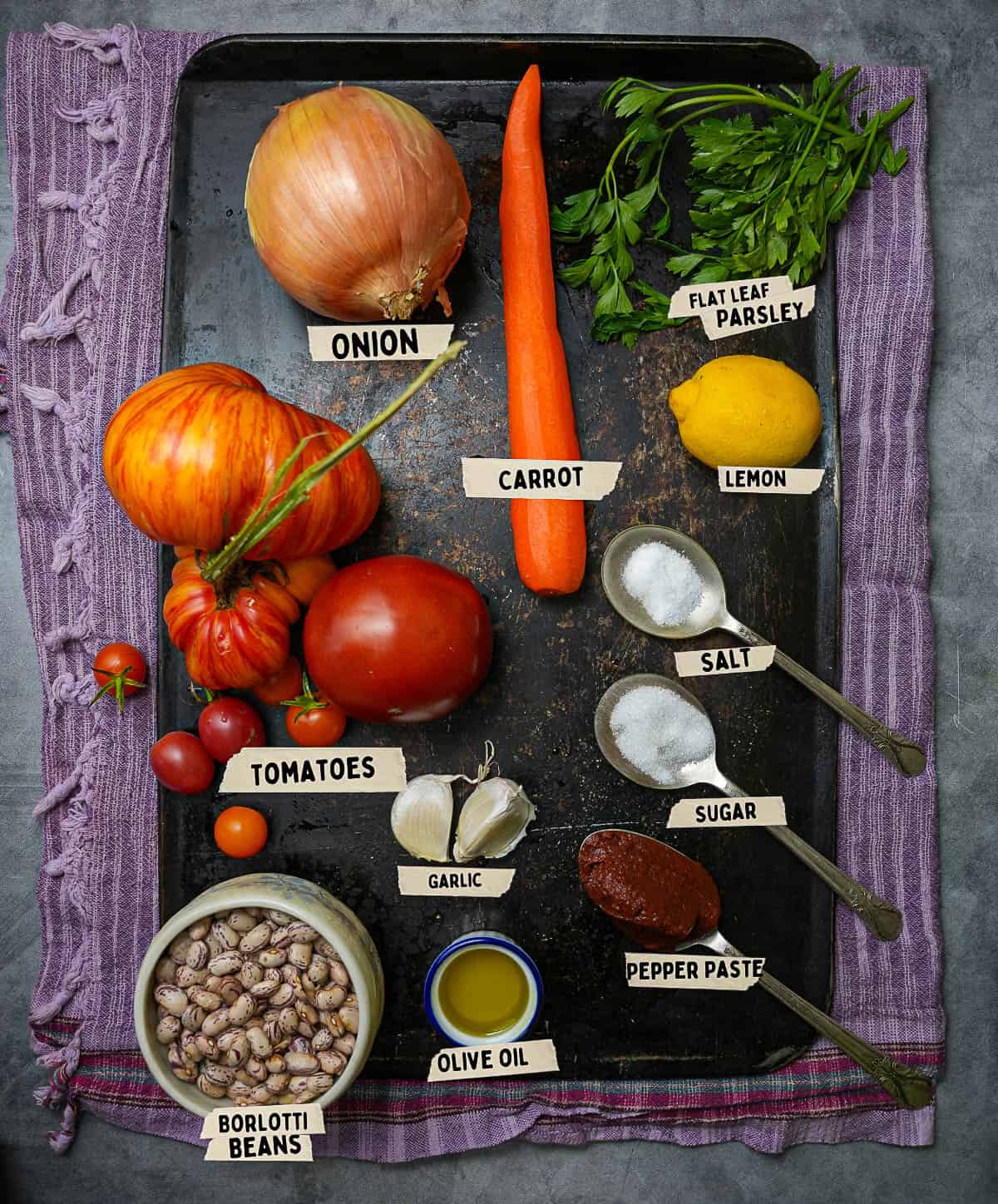 Ingredients for making Barbunya Pilaki are measured out and labeled on a dark metal tray.